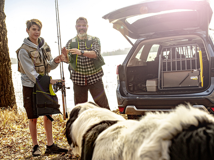 En man och en pojke plockar fram fiskeutrustning ur bakluckan på sin Subaru Forester, medan deras stora lurviga hund sitter i förgrunden – redo för en dag vid sjön.