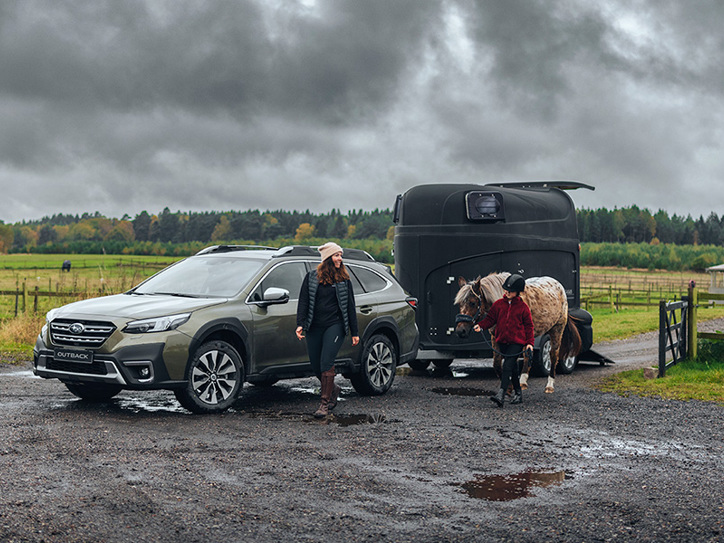 Subaru Outback med hästtrailer i lera, medan en kvinna och en flicka går bredvid – flickan leder en häst.
