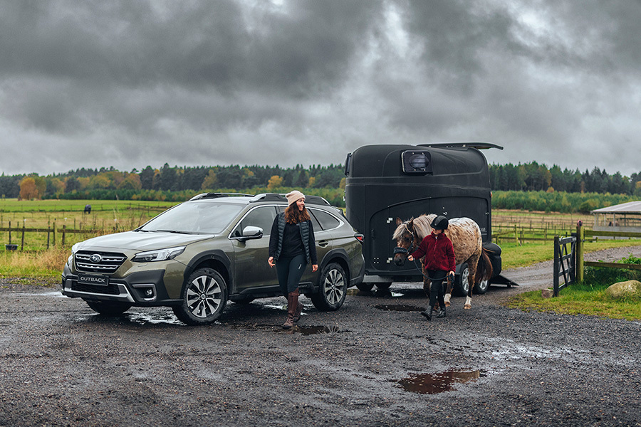 Subaru Outback med hästtrailer i lera, medan en kvinna och en flicka går bredvid – flickan leder en häst.